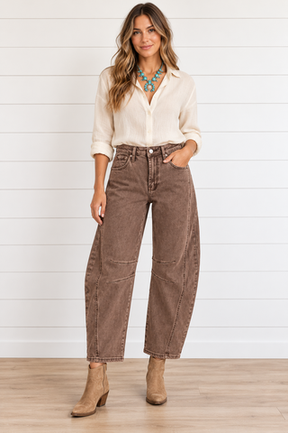 Model wearing washed brown barrel jeans with curved seams and knee darts, a cream blouse, turquoise necklace, and tan ankle boots.