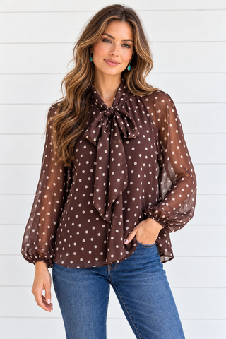 Female model wearing a chocolate brown and ivory polka dot bow-neck blouse with sheer long sleeves and blue jeans.