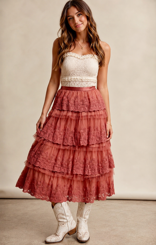 Model wearing a rust-colored tiered lace midi skirt with ruffle detailing, styled with an ivory lace top and white embroidered western cowboy boots.