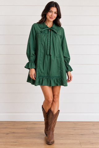 Female model wearing a forest green mini dress with a lace-trimmed collar, neckline bow, and ruffle cuffs, styled with brown cowboy boots.