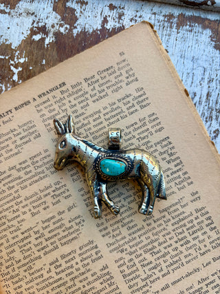  Antique-style silver donkey pendant featuring a turquoise stone accent, photographed on an open vintage book with a distressed wood background.