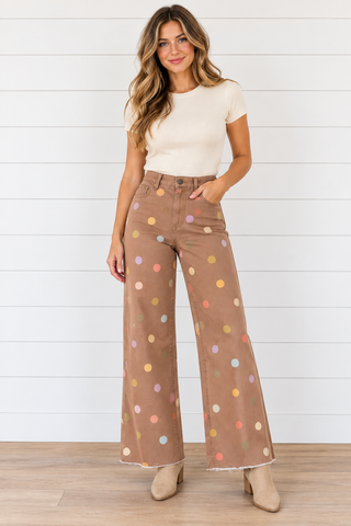 Female model wearing brown wide leg pants with multicolored polka dots and raw hems, styled with a cream top and beige boots.