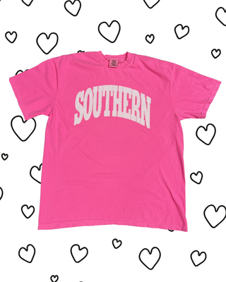 A bright pink short-sleeve t-shirt with 'SOUTHERN' printed in white arched block letters across the chest, displayed flat against a white background decorated with hand-drawn black heart outlines.