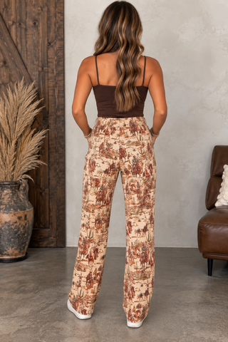 A woman shown from the back wearing high-waisted wide-leg pants with a brown and beige toile print, paired with a dark brown camisole top. She is standing in a styled room with a textured gray wall, a wooden door, a pampas grass vase, and a brown leather armchair.