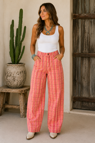 A woman stands in a rustic indoor setting wearing wide-leg plaid pants in pink and orange, paired with a white tank top and beige boots. She is posed next to a potted cactus and a weathered wooden door.