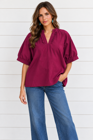 Model wearing a deep berry ruffle-neck blouse with elbow-length sleeves and blue jeans against a white shiplap wall.