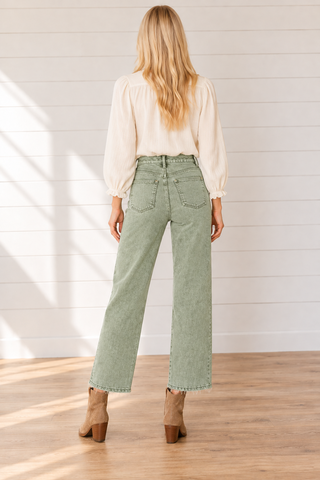 Back view of a model wearing washed olive wide-leg cropped jeans with two back pockets and raw hems, a cream blouse, and tan ankle boots against a white shiplap wall.