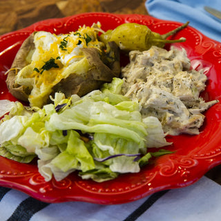 Crockstar Creamy Pepperoncini Chicken served with a loaded baked potato, green salad, and whole pepperoncini peppers on a red plate.