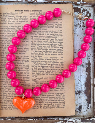Hot pink beaded necklace with an orange heart pendant and gold-tone lobster clasp, displayed on a vintage book page.