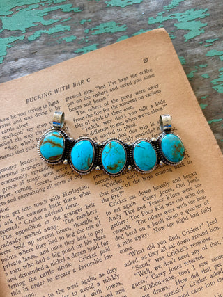 Curved bar pendant with five oval turquoise-colored stones, rope-style borders, silver-tone bead accents, and two bails.