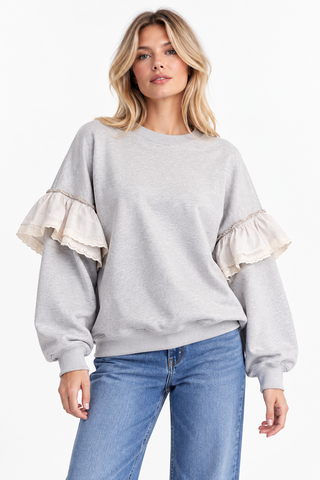 Model wearing a heather gray pullover with cream layered ruffle sleeves and blue jeans against a white background.