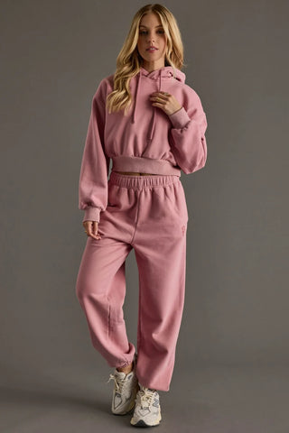 A woman modeling a dusty pink two-piece tracksuit consisting of a cropped pullover hoodie and matching sweatpants, paired with white sneakers, against a solid gray studio background.