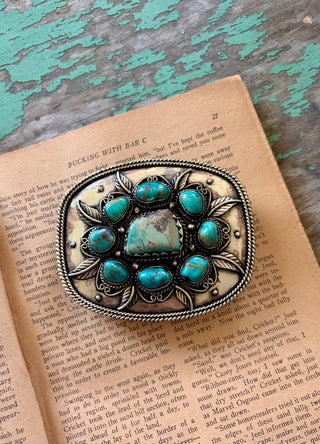 Antique silver-tone belt buckle with a large turquoise-colored center stone, eight surrounding stones, leaf accents, and a twisted rope border.