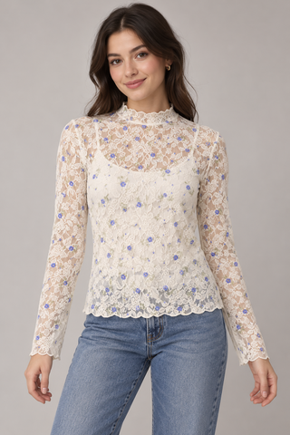 Model wearing a sheer ivory lace top with small blue flowers, a mock neckline, long sleeves, and scalloped edges over an ivory camisole with jeans\.