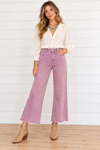 Model wearing dusty violet high-rise wide-leg cropped jeans with raw hems, a cream blouse, turquoise necklace, and tan boots.