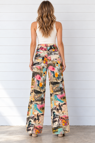 Model wearing full-length wide leg pants with a colorful cowgirl, horse, desert, and floral collage print, paired with a white tank and cream boots.