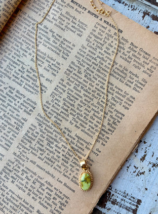 Oval yellow\-green Palomino turquoise pendant with a beaded gold border on a gold\-plated sterling silver chain, displayed on an open book\.