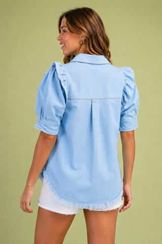 A woman shown from the back wearing a light blue chambray puff-sleeve blouse and white shorts against a solid olive green background.