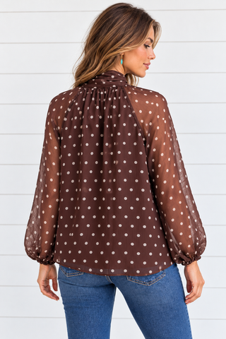 Back view of a chocolate brown and ivory polka dot blouse showing the gathered neckline, flowy hem, and sheer long sleeves.