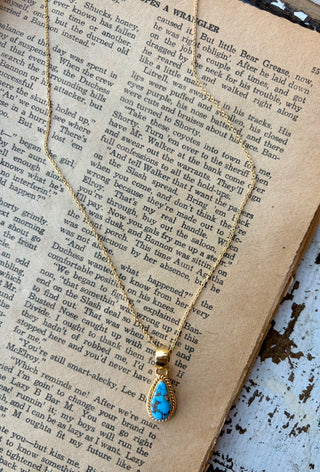 Blue Golden Hills turquoise teardrop pendant with dark veining and a gold rope-style border on a gold-plated sterling silver chain, displayed on an open book.