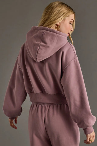 A woman shown from the back wearing a dusty mauve two-piece loungewear set consisting of a cropped hooded sweatshirt and matching sweatpants, against a solid gray studio background.