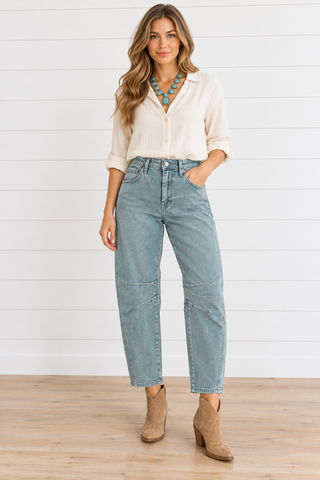 Model wearing washed teal barrel jeans with curved seams and knee darts, a cream blouse, turquoise necklace, and tan ankle boots.