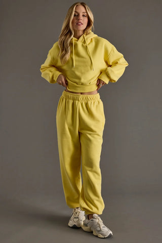 A woman modeling a matching two-piece lounge set in bright yellow, featuring a cropped long-sleeve hoodie and matching sweatpants, paired with white sneakers against a solid gray studio background.