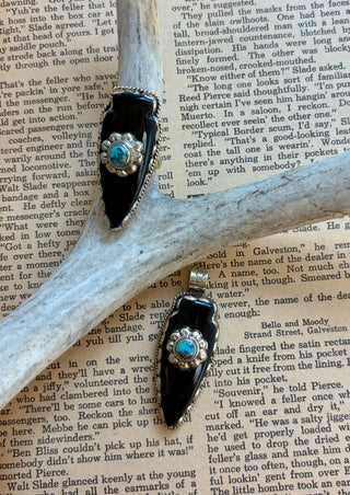 Handcrafted black onyx arrowhead pendant with a genuine turquoise accent set in an antique silver alloy setting, displayed on a vintage book background.