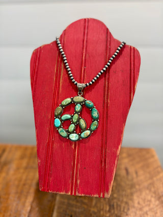 Turquoise peace sign pendant necklace featuring multiple oval turquoise stones with silver-tone detailing on a beaded necklace, displayed on a rustic red wooden jewelry stand.
