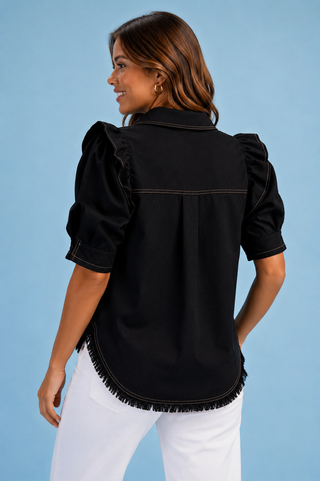 A woman wearing a black short-sleeve blouse with puffed sleeves, contrast stitching, and fringe trim along the curved hem, paired with white pants against a solid light blue background.