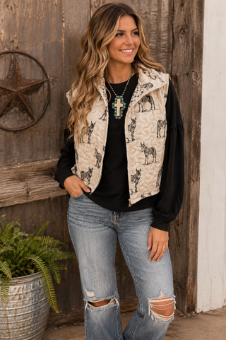 A woman wearing a cream quilted vest with black donkey illustrations, a black long-sleeve top, and distressed blue jeans, standing in front of a rustic wooden door.