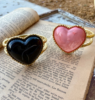 Pink and black oversized heart bracelets with beaded gold\-tone borders and textured double bands, displayed on an open book\.