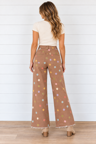 Female model wearing brown wide leg pants with multicolored polka dots and raw hems, styled with a cream top and beige boots.