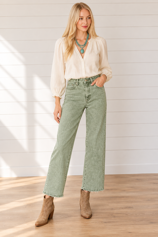 Model wearing washed olive high-rise wide-leg cropped jeans with raw hems, a cream blouse, turquoise necklace, and tan ankle boots against a white shiplap wall.