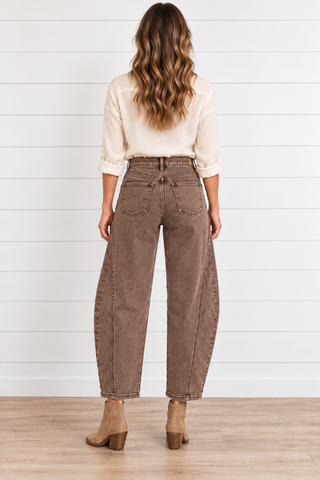 Back view of a model wearing washed brown barrel jeans with two patch pockets and cropped finished hems, a cream blouse, and tan boots.