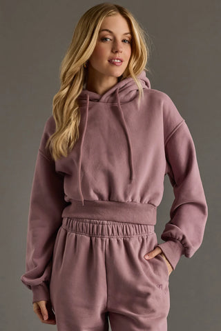 A woman modeling a matching dusty rose pink loungewear set, featuring a cropped pullover hoodie with drawstrings and matching jogger pants, set against a solid gray studio background.