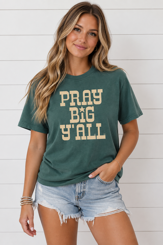 Woman wearing green Pray Big Y’all graphic tee with cream lettering and denim shorts