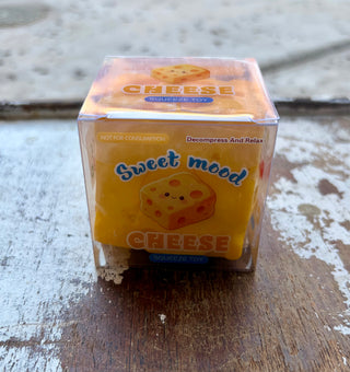 Yellow cheese-shaped squishy toy in a clear box labeled “Cheese Squeeze Toy,” sitting on a distressed wooden surface.