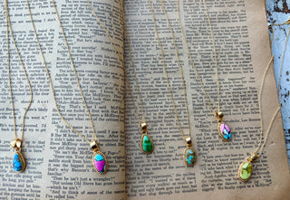 Six gold-plated sterling silver necklaces with blue, green, yellow-green, and multicolored stone pendants arranged across an open book.