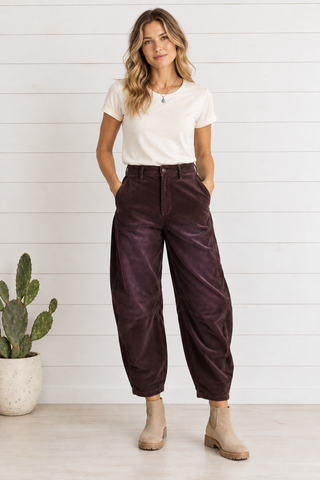 Female model wearing chocolate wine corduroy barrel pants with an ivory tee and beige boots against white shiplap beside a potted cactus.