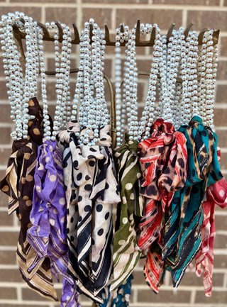 Pearl-style necklaces with thin scarves tied in front, featuring colorful polka dot and striped prints, hanging on a metal display rack.
