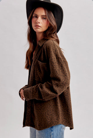 A woman wearing a brown leopard-print button-up shirt, blue jeans, and a black wide-brim hat, photographed against a light gray studio background.