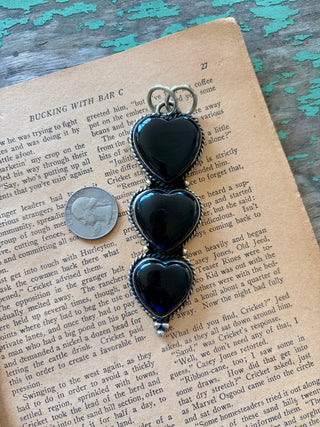 Three black onyx heart stones stacked vertically in a silver-tone alloy pendant with textured borders and decorative bead details.