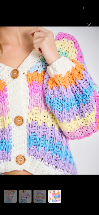 A close-up of a person wearing a chunky knit cardigan with horizontal pastel stripes in cream, pink, orange, blue, purple, and yellow, featuring three wooden buttons.