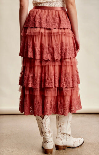 A tiered dusty rose lace and mesh maxi skirt with white western cowboy boots, shown from the back on a model.