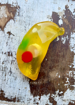 Translucent yellow jumbo banana squishy with colorful fruit-shaped pieces inside, displayed in a clear box on distressed white wood.