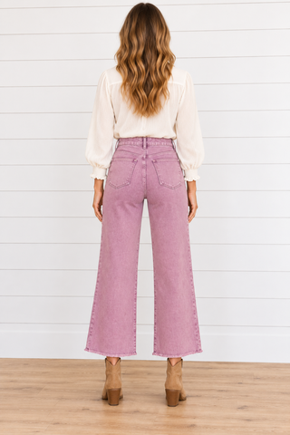 Back view of a model wearing dusty violet wide-leg cropped jeans with two patch pockets and raw hems, a cream blouse, and tan boots.