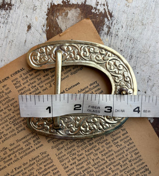 An ornate vintage brass buckle with intricate scrollwork, shown with a measuring tape to indicate its size, resting on a piece of antique book paper over a distressed wood background.