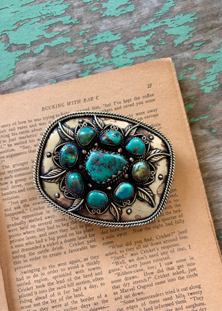 Antique silver-tone belt buckle with a large turquoise-colored center stone, eight surrounding stones, leaf accents, and a twisted rope border.