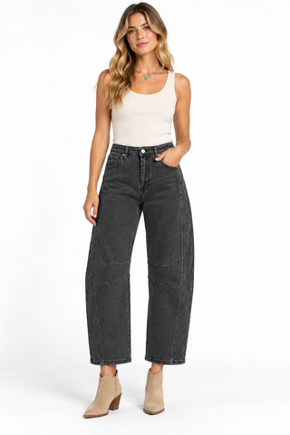 Model wearing washed black barrel jeans with curved knee seams, a cream tank top, and tan ankle boots.